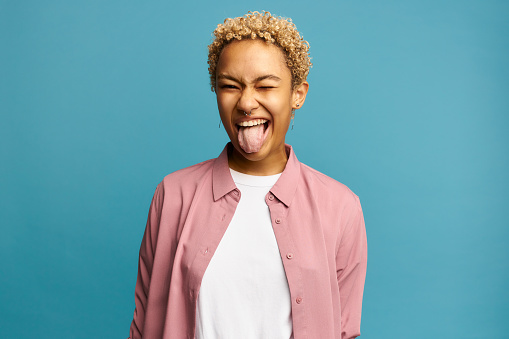 Pretty joyful carefree blonde African American girl with cute short curls, making faces in front of camera showing her tongue, dressed in pink shirt, having cheerful look, posing against blue wall
