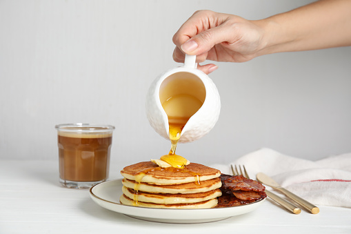 Woman pouring maple syrup onto delicious pancakes with butter and fried bacon at white wooden table, closeup