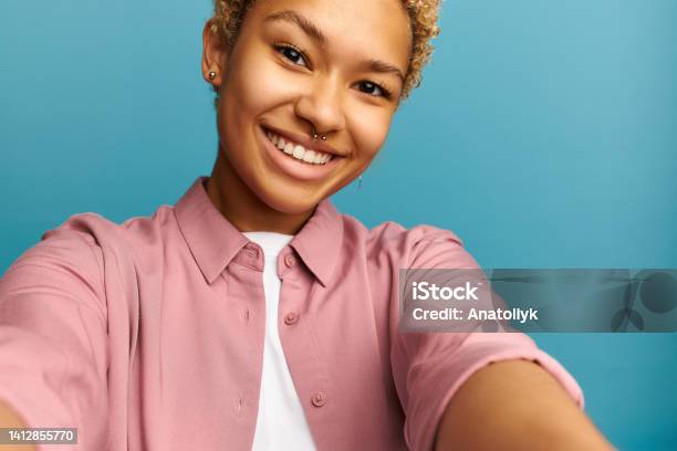 Crop shot of cheerful carefree blonde African girl taking selfie smiling widely, wearing nose ring, dressed in pink, isolated against blue background. Leisure time activity. Youth and technology