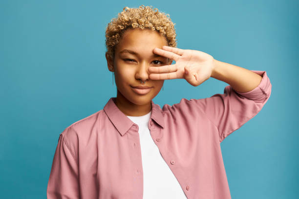 Beautiful pretty gorgeous blonde African American girl showing v-sign or peace and victory, looking through it closing one eye, wearing casual clothes standing against blue wall background Beautiful pretty gorgeous blonde African American girl showing v-sign or peace and victory, looking through it closing one eye, wearing casual clothes standing against blue wall background vulcan salute stock pictures, royalty-free photos & images