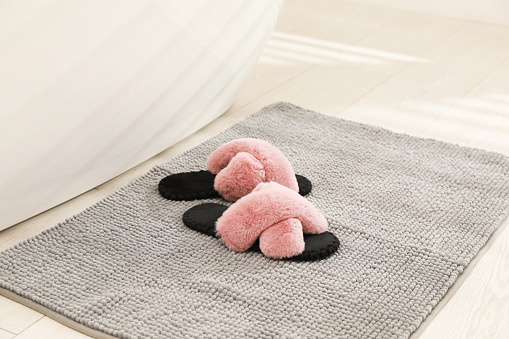 New grey bath mat with fluffy slippers near tub indoors