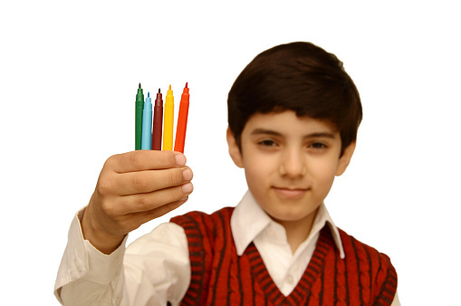 Cut out of a horizontal photograph of one confident schoolboy, looking at the camera and showing a bunch of coloured pens held tight raised isolated over white background. There is no text, and copy space for text. He has a mysterious smile and expressive eyes facial expressions. Focus is on hand and pens.