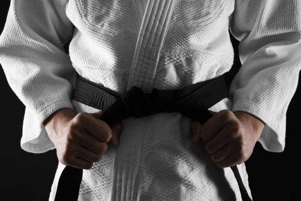 hombre en keikogi atando cinturón negro sobre fondo oscuro, primer plano. uniforme de artes marciales - judo fotografías e imágenes de stock