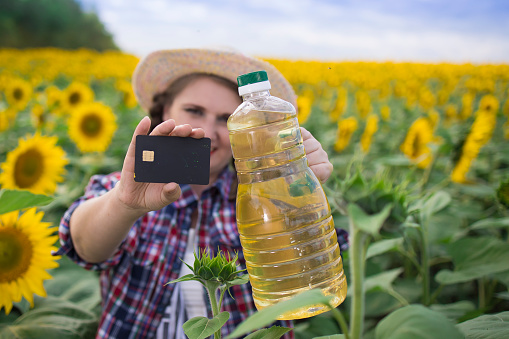 Beautiful smiling joyful middle-aged farmer woman  in a straw hat and a plaid shirt stands in defocus blur a field of  golden sunflower oil in her hands and  bank card for purchase in a harvest field of sunflowers on a sunny day