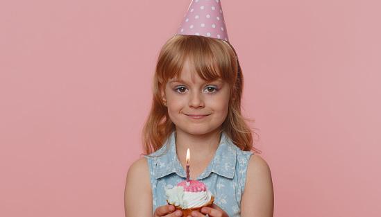 Happy young preteen child girl kid wears festive birthday hat hold cupcake makes wish joyful congratulating blowing burning candle on cake. Little toddler children celebrating anniversary party alone