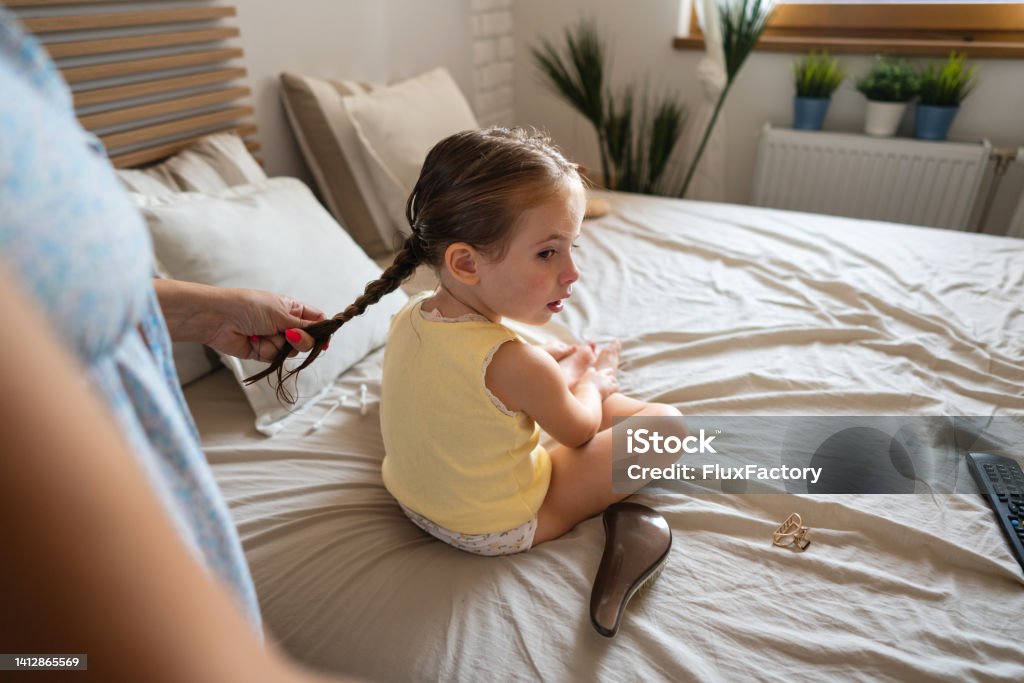 Unrecognizable mother making pigtails to her toddler daughter Caucasian toddler girl sitting on the bed while her mother making her pigtails Child Stock Photo
