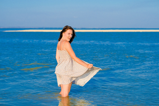 A young girl in a sundress fluttering in the wind stands knee-deep in the sea.