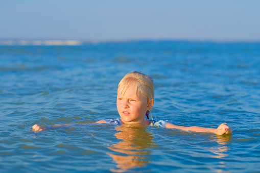 A blonde child girl bathes in the sea on a sunny summer day. The head of a happy child sticks out of the water.