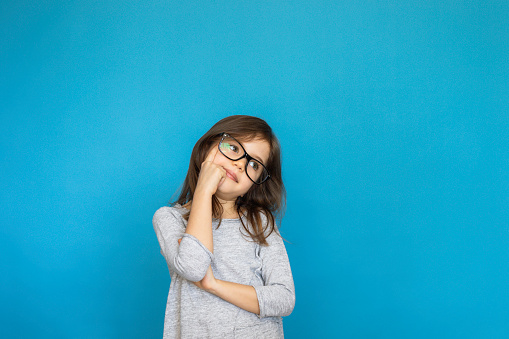 Little girl with eyeglasses is looking up with thinking gesture in front of blue background