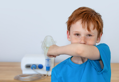 The boy coughs into his elbow, there is a nebulizer for inhalation behind him. The concept of medicine, healthcare, treatment of covid19