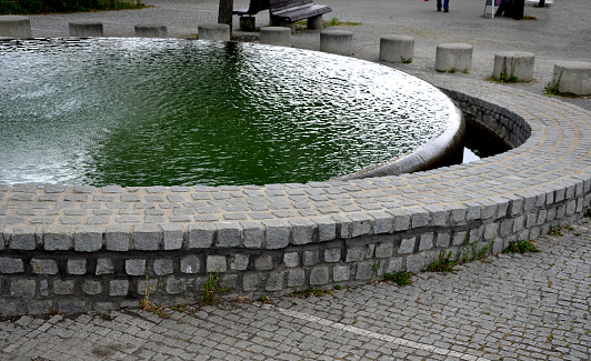 circular fountain with a level like a mirror up to the top of the edge of the wall. water flowing over tiled cobblestone granite wall. seat made of concrete cylinder bench. circle around the fountain.