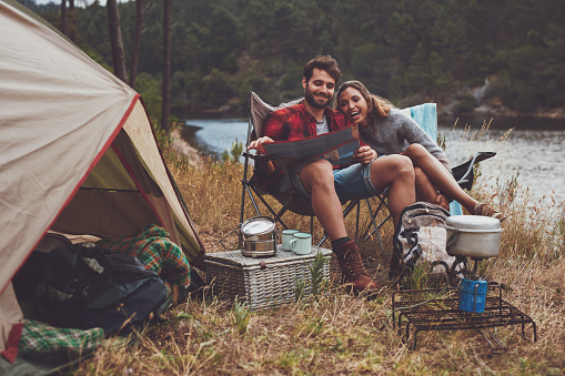 Loving young couple reading a travel map while sitting together outside their tent. Happy young couple camping next to the lake.