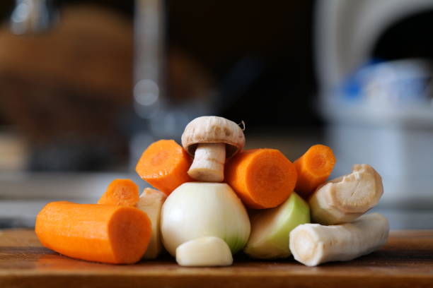 On the picture there is a bunch of cleaned vegetables (mushroom, onion, carrot, parsley, kohlrabi, celery, garlic) prepared for a soup lying on a wooden chopping board in the kitchen of a home. In the background we see the sink and other things related to dishwashing.