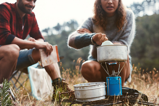 Young woman preparing food on a gas stove while camping with her boyfriend. Adventurous young couple camping in the forest.