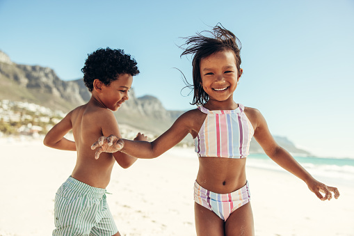 Two cheerful little kids having fun and enjoying themselves at the beach. Two adorable ethnic kids smiling happily while playing around in swimwear.