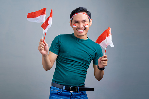 Portrait of cheerful young Asian man holding indonesian flag isolated on grey background. indonesian independence day on 17 august concept