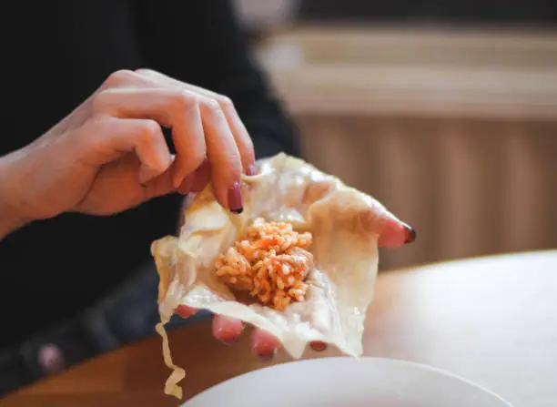 The hands of a young woman hold a straightened leaf of sauerkraut with rice-meat filling in the palm of her hand, folding it into a cabbage roll, sitting at a round table in the kitchen, close-up side view. The concept of step by step instructions, home cooking, traditional recipes, national cuisine, cabbage rolls, dolma.