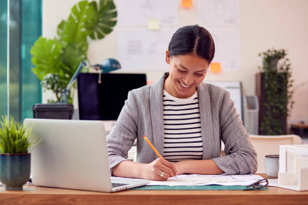 istock Female Architect Working In Office Sitting At Desk Studying Plans For New Building 1412883628