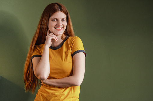 Smiling girl in yellow T-shirt standing in front green background and holding chin. She is looking at camera and smiling.