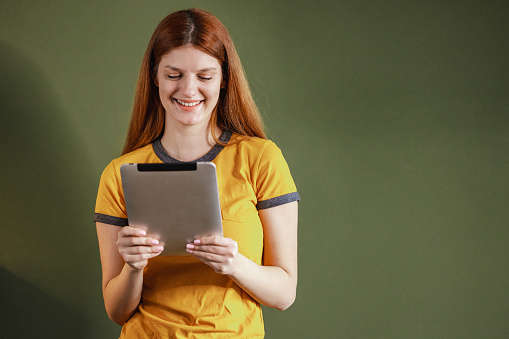 Shot  of a young woman holding digital tablet in her hands and reading data while standing against green background.