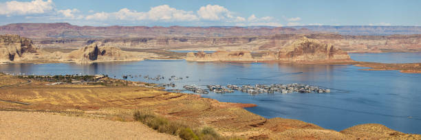 A wide panoramic view of Lake Powell at sunset, with houseboats and small boats docked at the marina. The sky transitions from warm orange near the horizon to a clear evening blue, with a few scattered clouds and the moon faintly visible. The desert landscape, red rocks, and sparse vegetation frame the shoreline, capturing the unique beauty of the American Southwest.