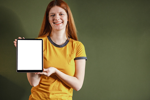 Shot of smiling  girl standing in front green background holding tablet with blank screen, sowing device and looking at the camera. Copy space concept.