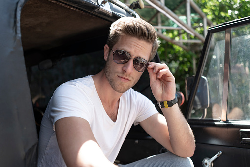 Handsome blond young man with stubble and sunglasses in front of black SUV.
