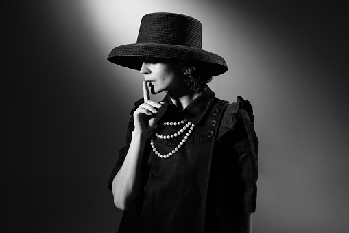 Portrait of beautiful woman in image of famous fashion designer posing in stylish black hat. Black and white photography. Secrets of fashion. Monochrome effect. Concept of fashion, style, history