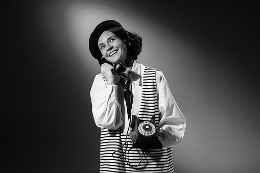 Portrait of beautiful emotive woman in stylish classical clothes talking on phone, smiling. Black and white photography. Cheerful communication. Monochrome effect. Concept of fashion, style, history