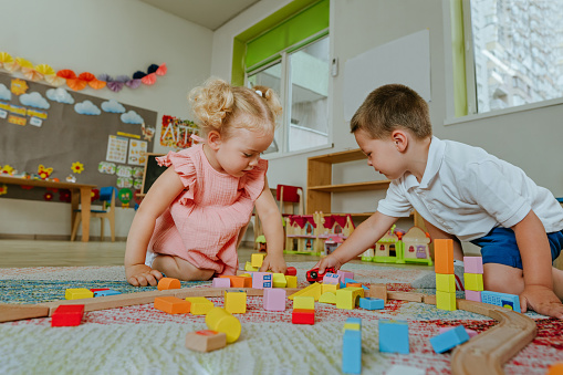 Toddlers boy and girl playing with wooden train sitting on the floor in kindergarten. Educational natural building blocks for kids.