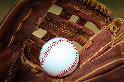 A close-up of a used yellow softball in a leather glove along with a pair of black cleats sitting in the grass