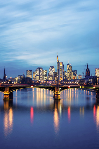 A long exposure vertical view of Frankfurt's skyline during the 'blue hour' following sunset, with the city lights reflected in the Main river.