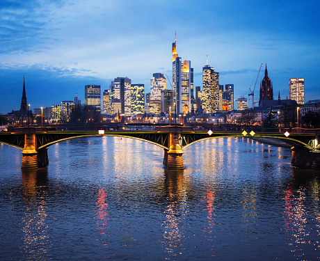 The towers of Frankfurt's financial district and the bridge over the River Main illuminated at dusk and reflecting in the water.