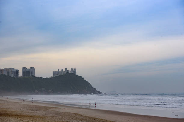 A wide shot captures a beautiful sandy beach with gentle waves rolling in, leading to a lush green hill on the right under a blue sky with wispy clouds.