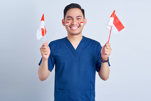 Handsome Asian man wearing blue male nurse uniform holding indonesian flag and celebrating indonesian independence day August 17 isolated on white background studio portrait