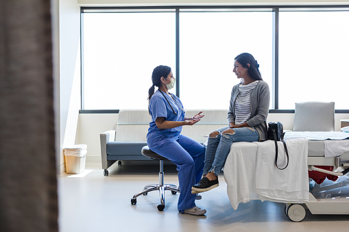The young adult female doctor gestures while talking to the young adult female patient in the ER examination room.