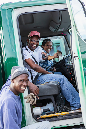 A multiracial group of three workers working at a lumberyard. An Hispanic woman and African-American man, in their 40s, are sitting in a truck, looking at the camera through the open door. The woman is the truck driver. The focus is on  the man. Their coworker is standing outside the truck.