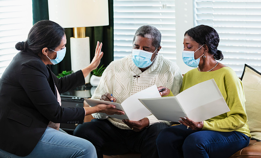 A senior African-American couple meeting with an in-home consultant. The mature Hispanic woman could be a Real Estate Agent, a financial advisor, or insurance agent. They are sitting in the living room signing documents. They are wearing protective face masks, trying to prevent the spread of coronavirus during the Covid-19 pandemic.