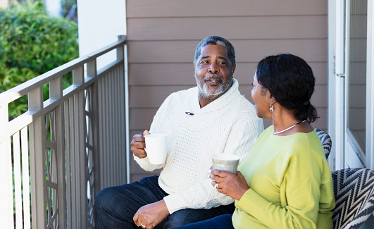 A senior African-American couple relaxing together on the terrace of their apartment. They are sitting on chairs, drinking coffee and conversing. The focus is on the man.
