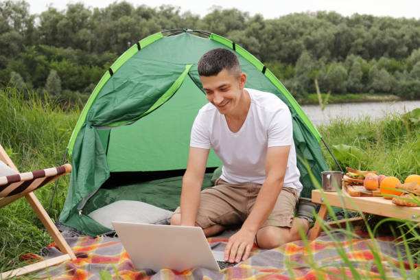istock Brunette male tourist working on a laptop outdoors in a camping, young guy freelancer working online at a laptop computer on a hike near rirver outdoors. The ability to work from anywhere in the world. 1412906578