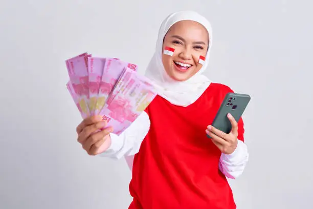 Smiling young Asian muslim woman in red white t-shirt holding mobile phone and cash money in Indonesian rupiah banknotes isolated on white background. Indonesian independence day on 17 august concept