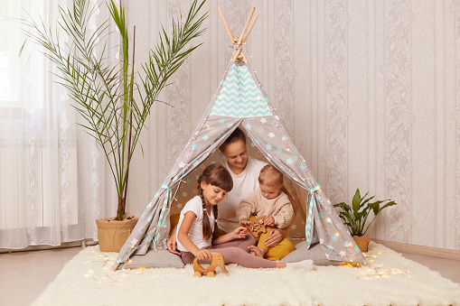 Image of Caucasian mother with her children sitting together teepee tent, mom playing with kids in play tent, mommy spending weekend with kids, playing and having fun in wigwam.