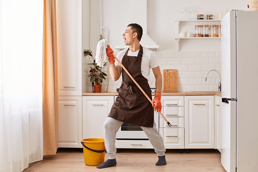 Full length photo of young guy cleaning kitchen, having fun, listening music and singing song, using mop as microphone, dancing, being in good mood while cleaning his apartment.