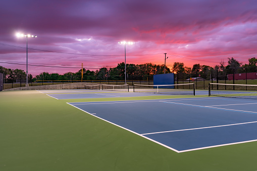 Evening photo of outdoor blue tennis courts with pickleball lines with lights turned on.