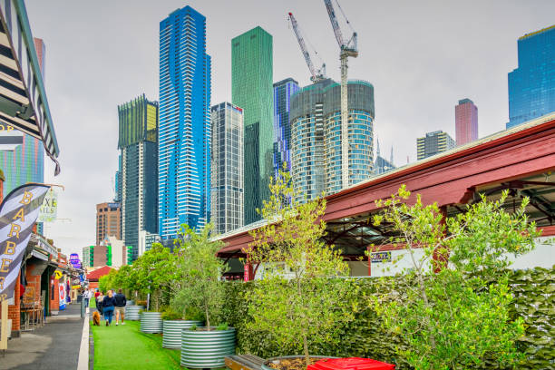 Pedestrians walk past stores at Queen Victoria Market in Melbourne, Victoria, Australia, with new condo towers in the background, on an overcast day.