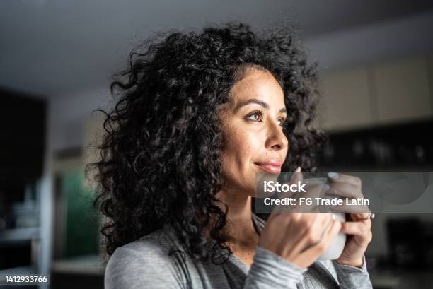 Mid Adult Woman Looking Away Holding Coffee Mug In The Kitchen At Home Stock Photo - Download Image Now
