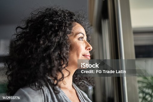 istock Mid adult woman looking through window at home 1412933776