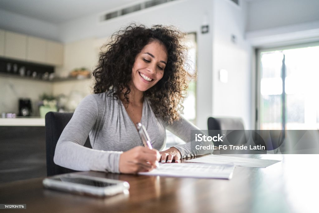 Mid adult woman filling document at home Mid adult woman filling paperwork at home Writing - Activity Stock Photo