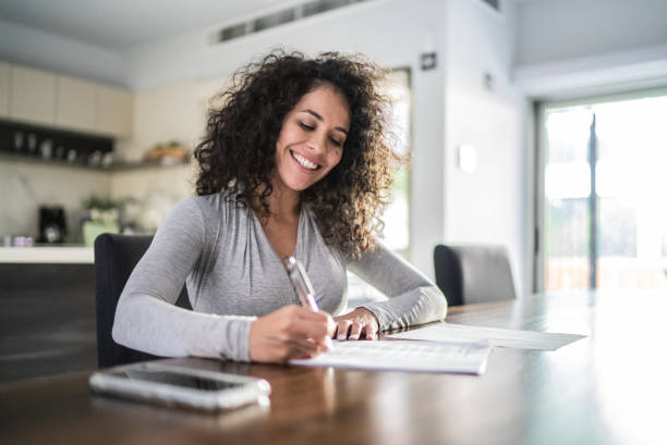 mid adult woman filling document at home - capital letter fotos imagens e fotografias de stock