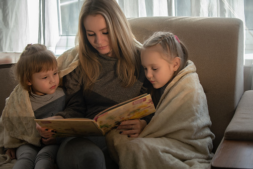 Mom at home on the couch reads a book to her little daughters. Family developing leisure at home